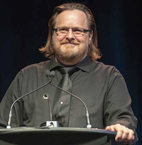 Marty Warren, Le directeur national du Syndicat des Métallos Un homme portant la barbe, des lunettes, une chemise noir et une cravate debout sur un podium s’adresse à une foule hors champ.