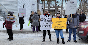 Un groupe de 15 personnes, la plupart des femmes, portant des tuques et de lourds manteaux d&rsquo;hiver, tiennent des pancartes, manifestant dans une rue de la ville. Le sol est enneig&eacute;.
