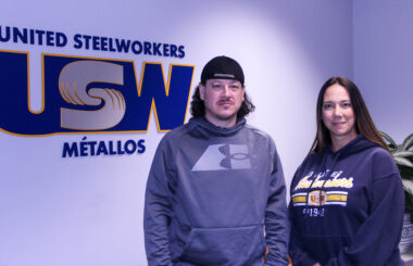 Image: A man and a woman wearing sweatshirts stand in a reception area facing the camera. Behind them is a giant USW logo on the wall of the space. 