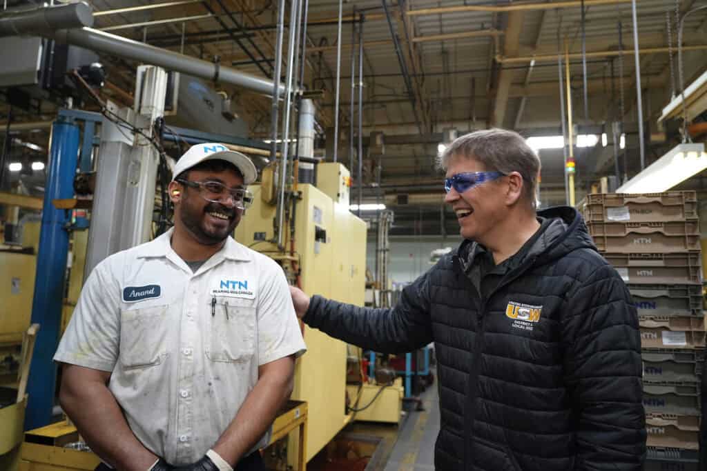 Image: a Black man and a white man greet each other, smiling, in an industrial setting. The Black man is wearing a work uniform with NTN on the