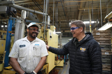 Image: a Black man and a white man greet each other, smiling, in an industrial setting. The Black man is wearing a work uniform with NTN on the 