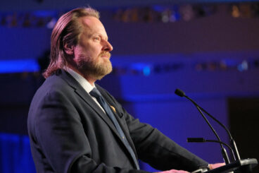 Image: profile image of a bearded man in a suit and tie standing at a podium with microphones looking out into an audience outside the frame. There is a wash of royal blue lighting in the background. 