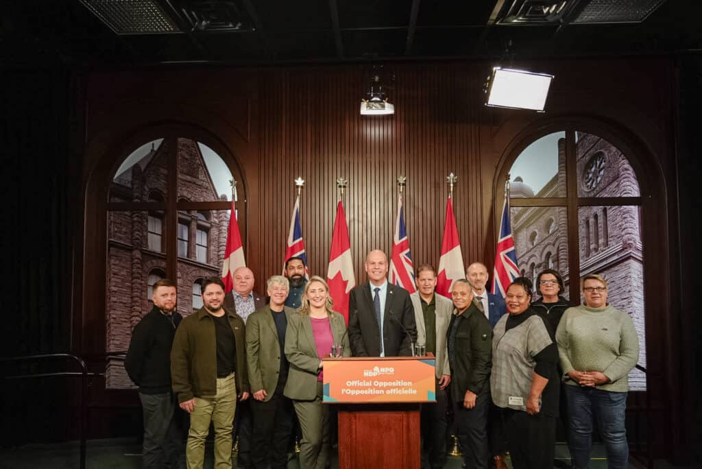 An image of a group of people standing side-by-side indoors. There is a podium in front of the person standing in the middle of the group. Behind them are flags