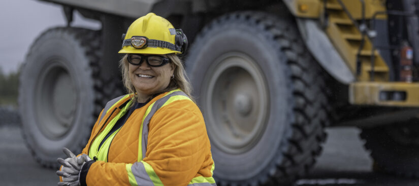 Photo: A woman wearing an orange safety vest and a hard hat is walking to the left of the frame. Behind her you can see part of a large heavy equipment truck used in mining.