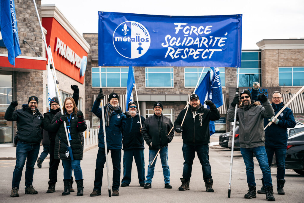 A group of Métallos at a picket, raising a flag with the slogan "Force, solidarité, respesct" [Eng.: Force, Solidarity, Respect]