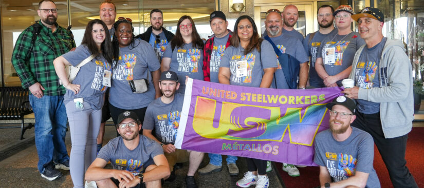 A photo of a big group of people gathered in front of hotel doors, outside. Most are standing side by side with a few sitting on the curb. There is a rainbow United Steelworkers USW Métallos flag being held in the middle .