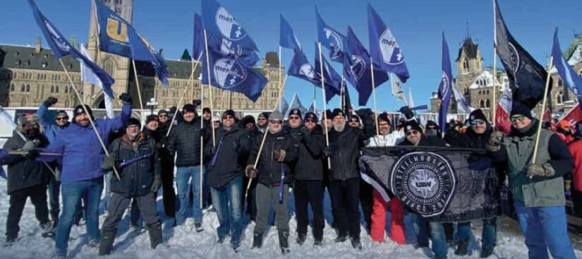 Des personnes sont prises en photo durant une manifestation à l’extérieur sur la Colline du Parlement. Elles portent des vêtements d’hiver et se tiennent debout sur la neige. Certaines brandissent des drapeaux bleus arborant divers logos du Syndicat des Métallos. L’édifice du Parlement canadien est visible en arrière-plan.