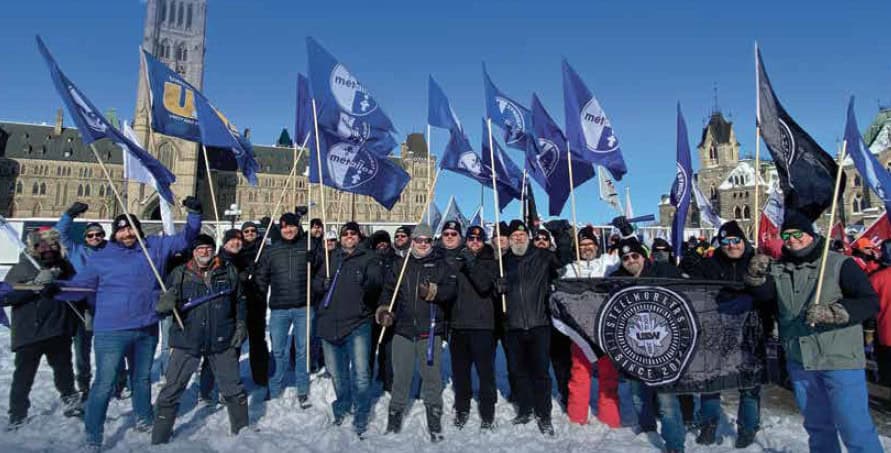 Des personnes sont prises en photo durant une manifestation à l’extérieur sur la Colline du Parlement. Elles portent des vêtements d’hiver et se tiennent debout sur la neige. Certaines brandissent des drapeaux bleus arborant divers logos du Syndicat des Métallos. L’édifice du Parlement canadien est visible en arrière-plan.