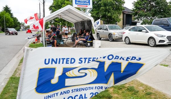 Image: A USW/Métallos Local 2918 flag on the foreground. In the background is a tent with  people sitting on chairs inside it.