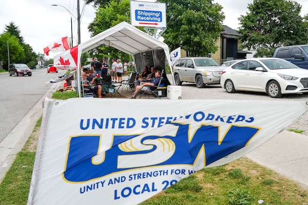 Image: A USW/Métallos Local 2918 flag on the foreground. In the background is a tent with  people sitting on chairs inside it.