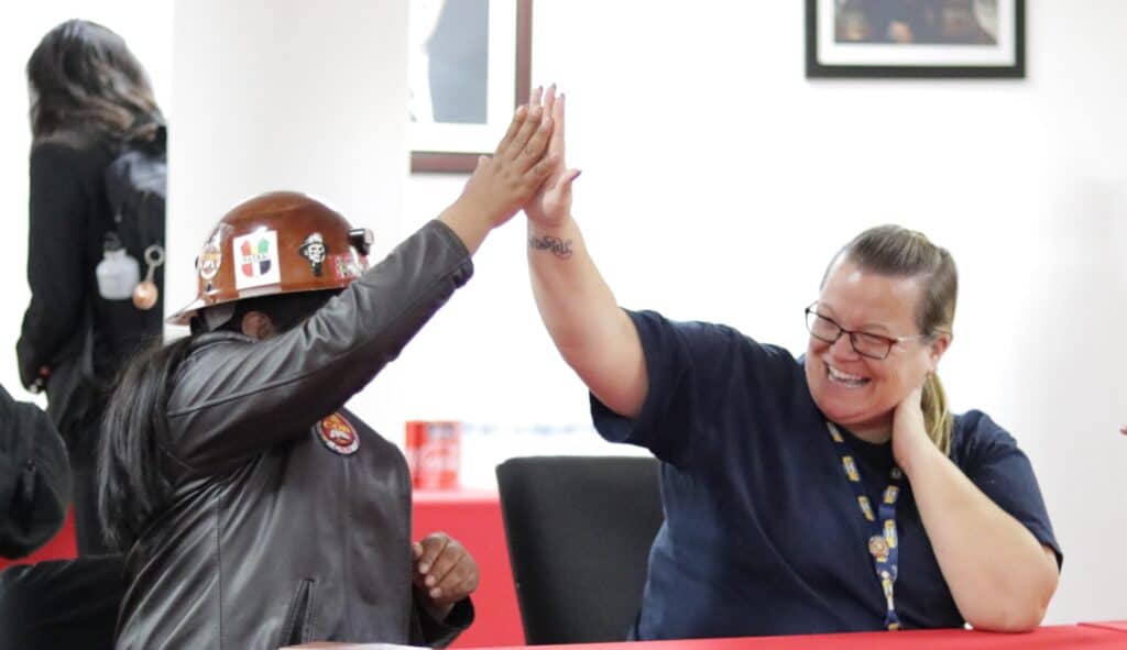 Two individuals sgiving each other a high five while sitting behind a table in a room. One of them is wearing safety helmet.