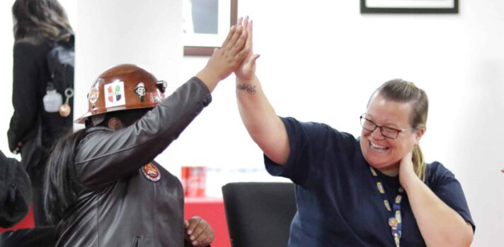 Two individuals sgiving each other a high five while sitting behind a table in a room. One of them is wearing safety helmet.