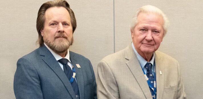 Two people in suit jackets stand side by side facing the camera with their hands folded in front of them. 