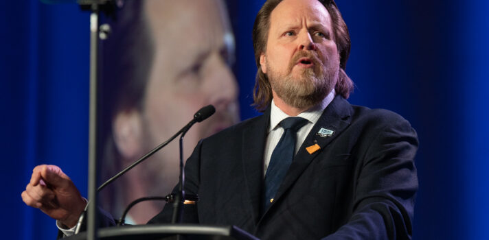 Image: man stands at microphone while speaking at a podium. They are dressed in a suit. 