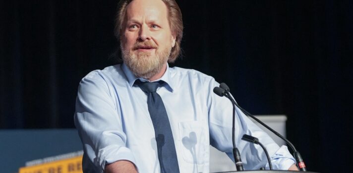 Image: man stands at microphone in front of large conference of 500 people.