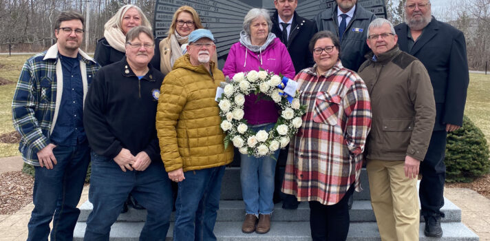 Un groupe de 11 personnes se tient en plein air devant un grand monument en granit. Au centre, l’un des membres du groupe tient une couronne de roses blanches.