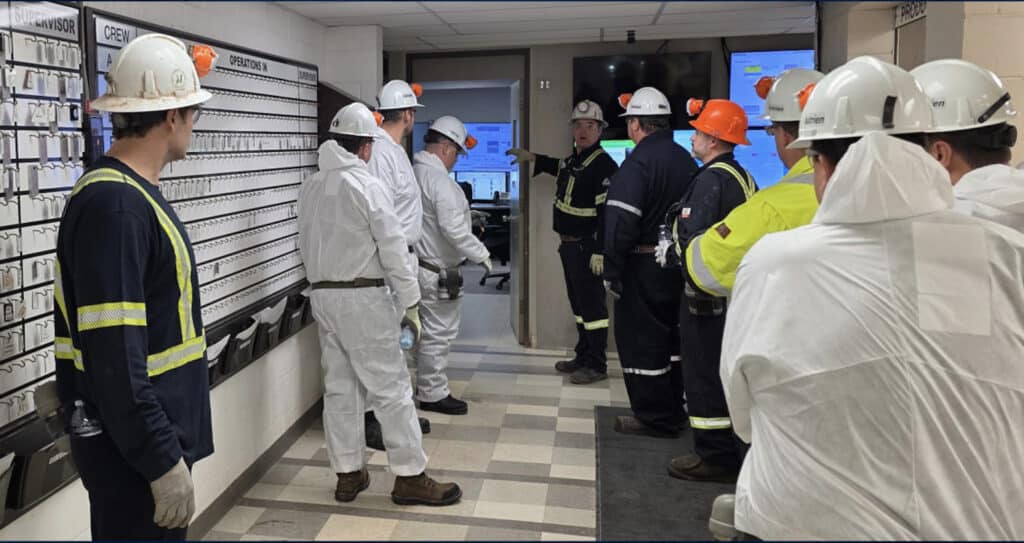 A group of ten workers in an interior space, facing away from the camera and towards a door to another room. They are in an industrial setting, wearing coveralls, some all navy and some all white, all wearing white hardhats with headlamps.