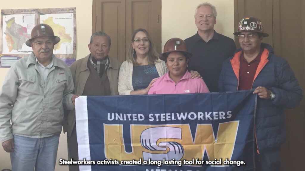 A group of workers from Latin America in an indoor setting, some wearing hardhats, holding a USW flag