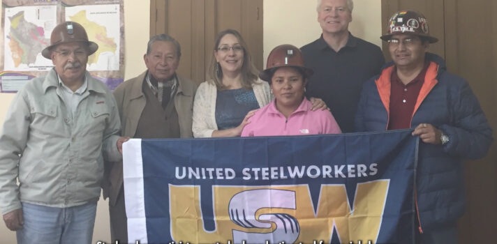 A group of workers from Latin America in an indoor setting, some wearing hardhats, holding a USW flag