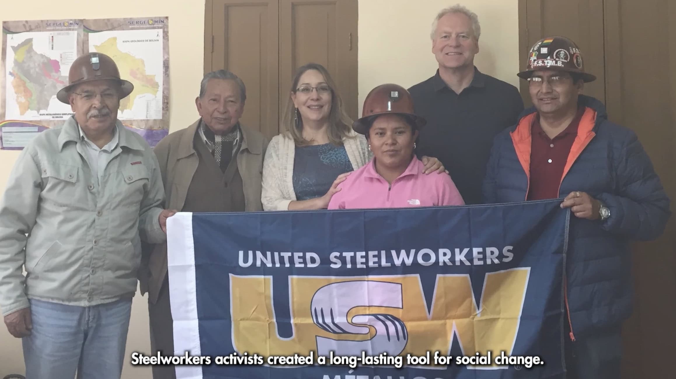 A group of workers from Latin America in an indoor setting, some wearing hardhats, holding a USW flag