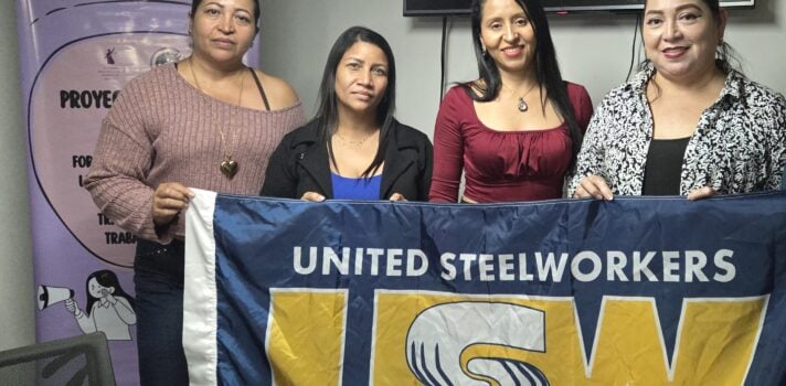 Four women stand together indoors holding a large United Steelworkers (USW) Métallos flag. A project banner is partially visible in the background.